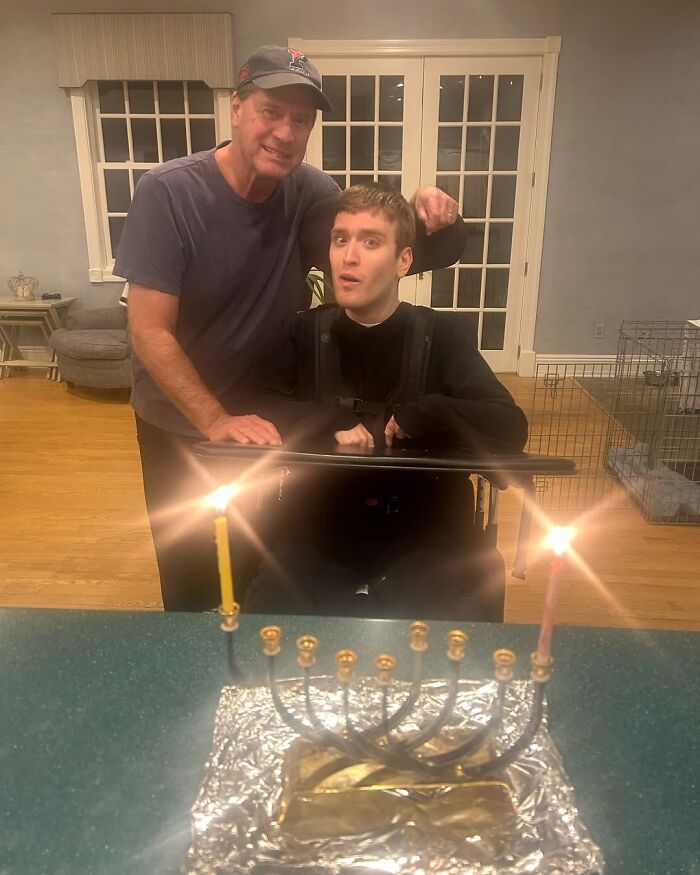 Man standing beside his disabled son in a wheelchair, with a lit menorah in the foreground, discussing Trump’s nephew.