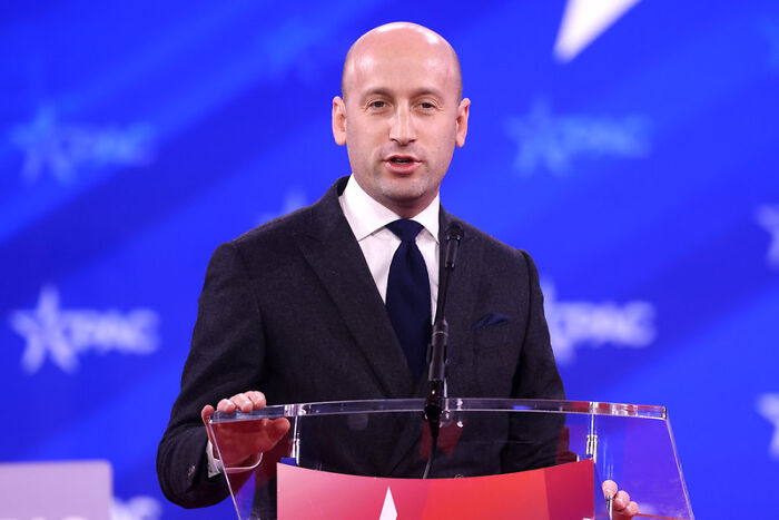 Man in suit speaking at a podium during a conference with a blue background featuring white star logos. Man in suit speaking at a podium during a conference with a blue background featuring white star logos.