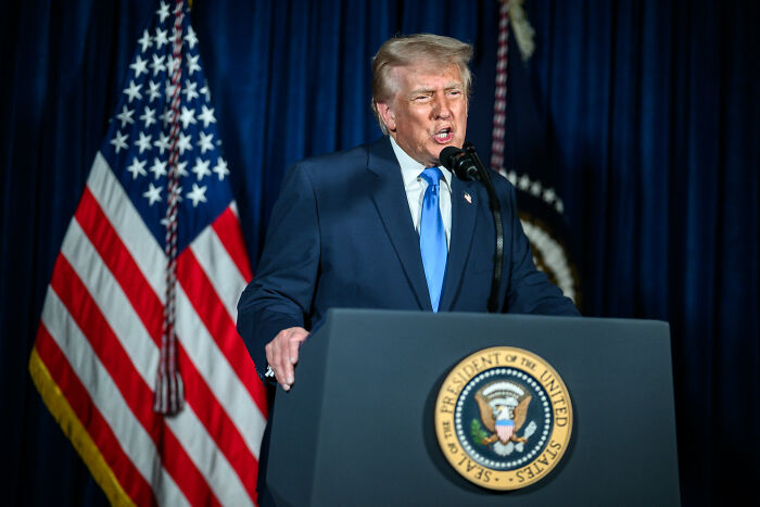 Former President Trump speaking at a podium with the presidential seal, flanked by American flags, amid MAGA base division. Former President Trump speaking at a podium with the presidential seal, flanked by American flags, amid MAGA base division.