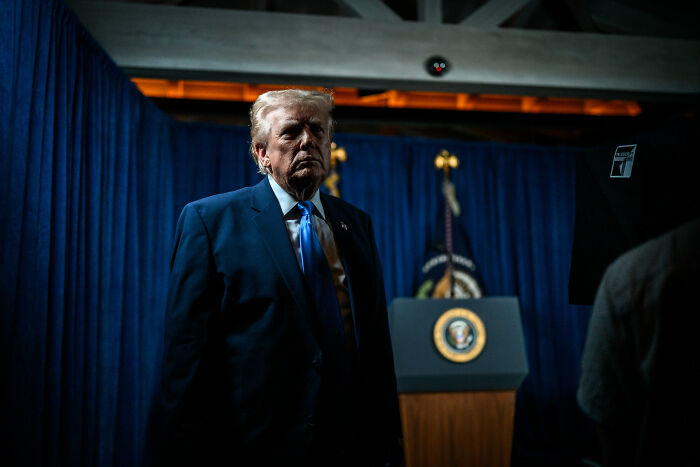 Donald Trump standing near a podium with the presidential seal, dimly lit against blue curtains. Donald Trump standing near a podium with the presidential seal, dimly lit against blue curtains.