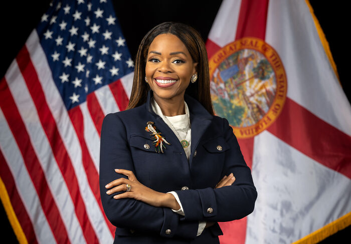 Lawmaker standing with arms crossed wearing a ring, smiling in front of US and Florida flags.