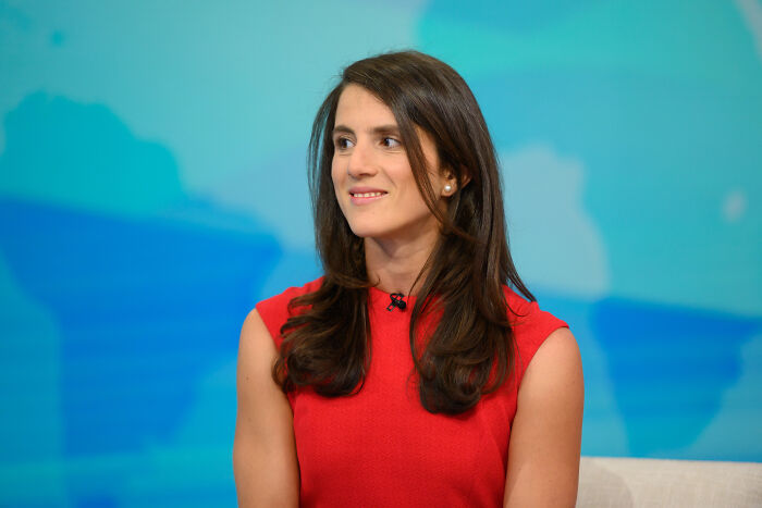 Woman in red dress seated in a studio setting, with a backdrop featuring abstract blue and green shapes, discussing Kennedy attacks.