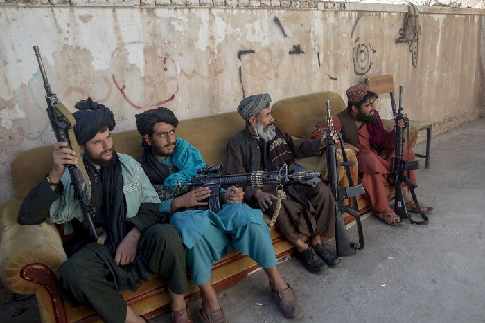 Four armed Taliban fighters sitting on a couch holding rifles against a wall in a desert setting. Four armed Taliban fighters sitting on a couch holding rifles against a wall in a desert setting.