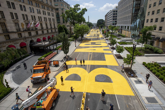 BLM Plaza in Washington DC with large yellow letters painted on the street and people walking along the area. BLM Plaza in Washington DC with large yellow letters painted on the street and people walking along the area.