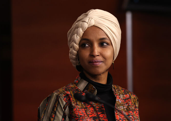 Ilhan Omar speaking at an event, wearing a beige headwrap and patterned jacket, highlighting ICE actions on Somali immigrants.