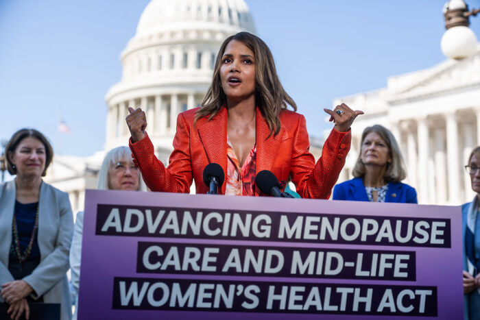 Halle Berry speaking at a podium about women’s health care, with the US Capitol building visible in the background.