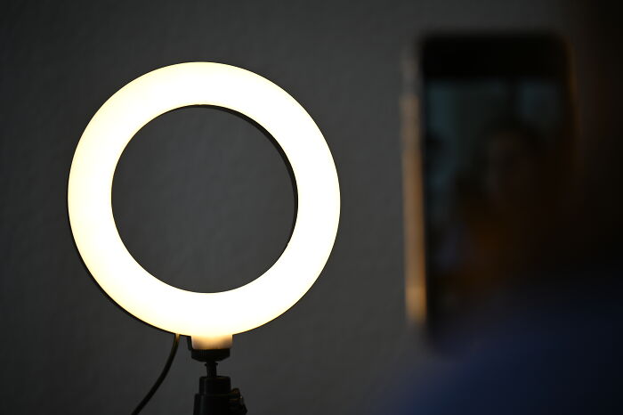 Ring light glowing brightly on a stand with a blurred person holding a smartphone nearby for recording or streaming. Ring light glowing brightly on a stand with a blurred person holding a smartphone nearby for recording or streaming.