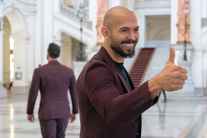 Man in a maroon suit smiling and giving thumbs up in a grand hall, related to misogynistic influencer and human trafficking topic.