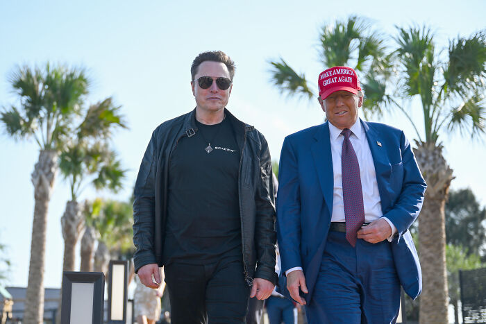Man in a black jacket and another in a suit with a red hat walking outdoors, related to political scandals 2025.