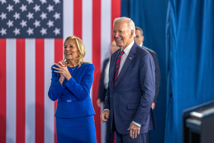 President Joe Biden and Jill Biden standing and smiling with an American flag backdrop during an event. President Joe Biden and Jill Biden standing and smiling with an American flag backdrop during an event.