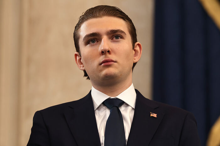 Barron Trump in a dark suit with an American flag pin, looking serious against a blurred background.