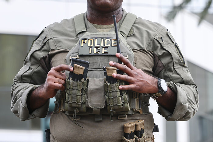 ICE police officer wearing tactical gear with visible POLICE ICE patch on vest outdoors in daylight.