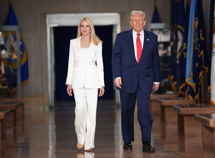 Pam Bondi walking alongside a man in a suit in a hallway with flags, appearing confident and formal. Pam Bondi walking alongside a man in a suit in a hallway with flags, appearing confident and formal.