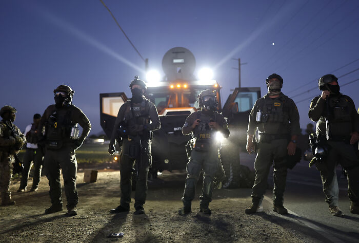 Heavily armed police officers stand in front of armored vehicle during Trump’s ICE raids showing arrested but not convicted criminals.