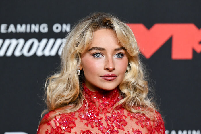 Sabrina Carpenter at an event, wearing a red lace dress, responding publicly to ICE using her song in anti-immigration video