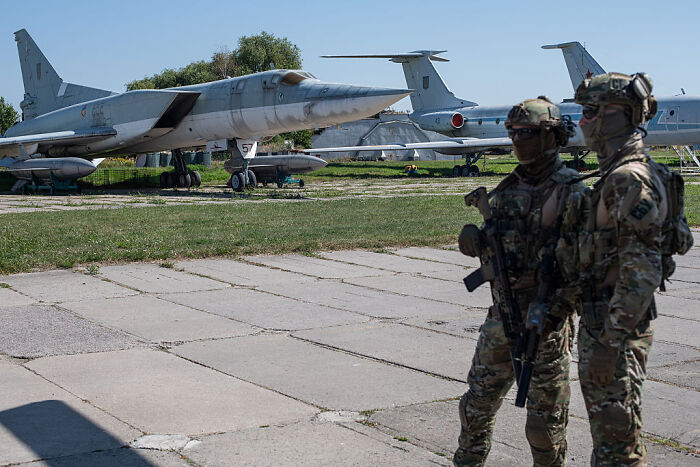 Two soldiers stand armed near military jets at an airfield related to hungover Russians impacting Ukraine’s deep strike plan.