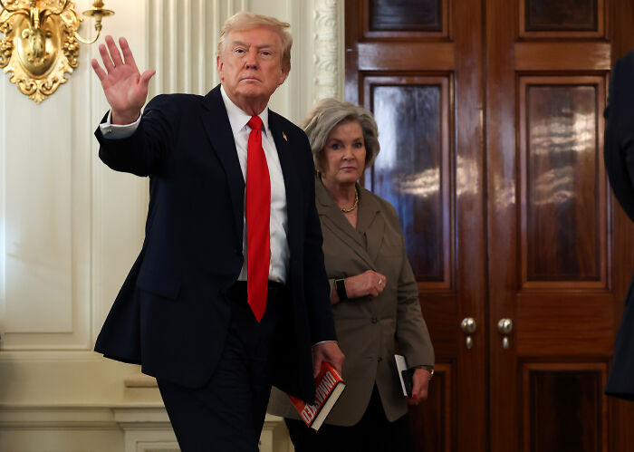 Donald Trump in a suit waving, with his chief of staff holding a notebook, inside a formal room with wooden doors. Donald Trump in a suit waving, with his chief of staff holding a notebook, inside a formal room with wooden doors.