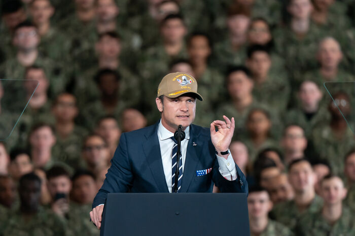 Pete Hegseth speaking at a podium in front of military personnel, wearing a suit and a tan cap with a logo.