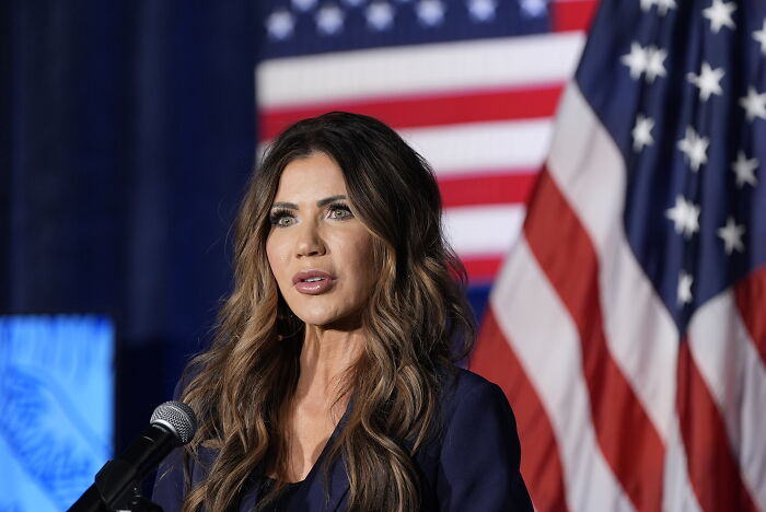 Kristi Noem speaking at event with American flags in background during hot mic moment involving TSA staff fear. Kristi Noem speaking at event with American flags in background during hot mic moment involving TSA staff fear.