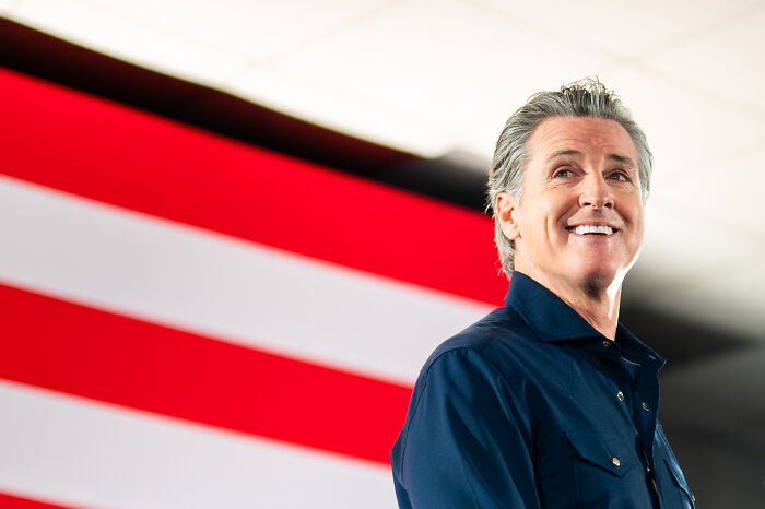 Gavin Newsom smiling in a dark shirt with an American flag backdrop highlighting exceptional bone density discussion.
