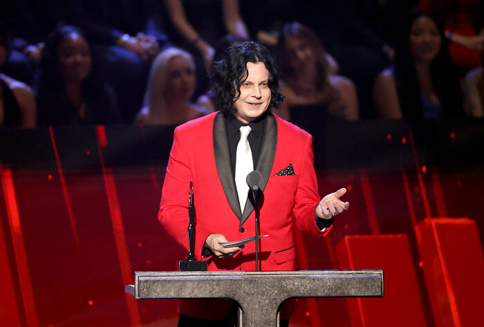 Jack White speaking at a podium in a red suit, addressing an audience during a live event.