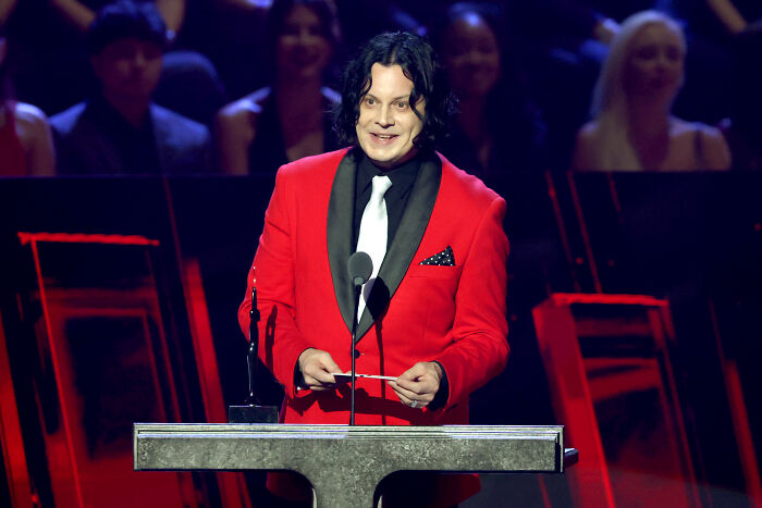 Musician in red jacket speaking at a podium with audience blurred in the background, related to celebrities slamming Donald Trump. Musician in red jacket speaking at a podium with audience blurred in the background, related to celebrities slamming Donald Trump.