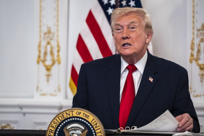 Former President Trump speaking at a podium with the American flag, linked to freeing GPB Capital founder jailed for fraud.