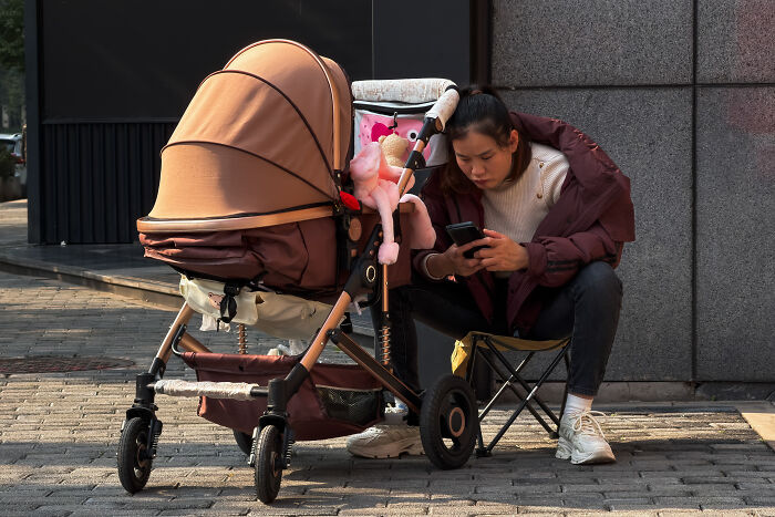 Mom sitting beside baby stroller outdoors, focused on phone, illustrating mom with baby in public setting.