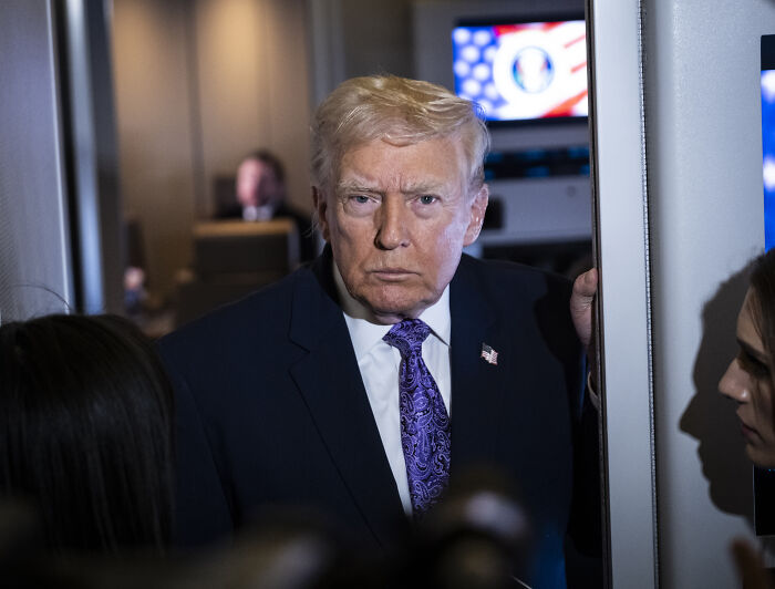 Trump in a dark suit and purple tie, appearing serious while interacting with reporters during an indoor event. Trump in a dark suit and purple tie, appearing serious while interacting with reporters during an indoor event.