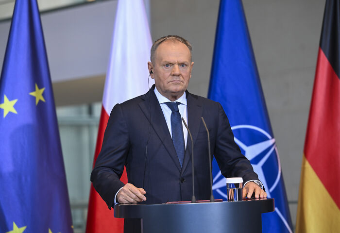 Man in dark suit standing at a podium with European Union, Poland, NATO, and Germany flags discussing terrifying events near WW3.