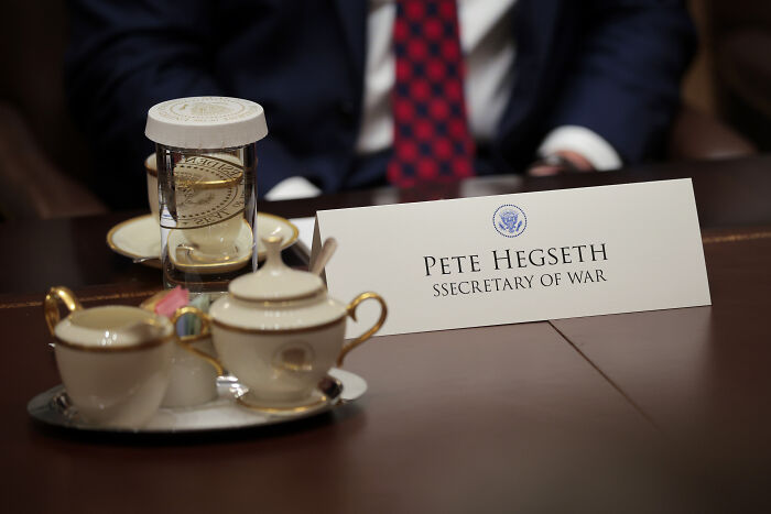 Nameplate for Pete Hegseth Secretary of War on a table during Trump’s cabinet meeting with tea set and hourglass nearby.
