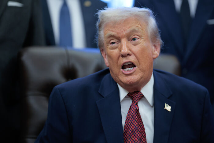 Former President Trump speaking in the Oval Office wearing a dark suit and red tie during a press event.