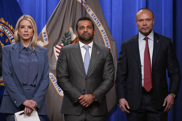 Three serious officials standing in front of Department of Justice flags during FBI pipe b**b investigation press briefing.