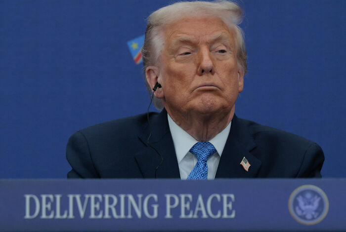 Donald Trump at a conference with an earpiece, in front of a sign about delivering peace, discussing renaming Kennedy Center.