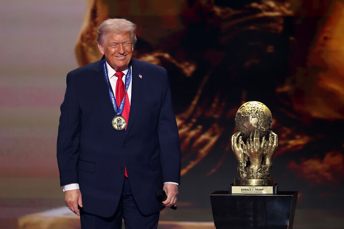 Donald Trump wearing a medal standing next to a gold trophy, related to FIFA ethics complaint and Infantino peace prize. Donald Trump wearing a medal standing next to a gold trophy, related to FIFA ethics complaint and Infantino peace prize.