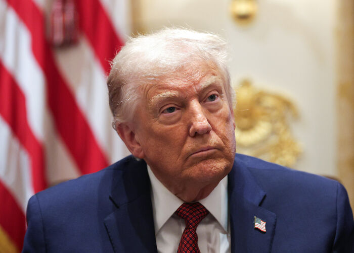 Donald Trump in a suit with an American flag pin, seated indoors with a serious expression and a blurred flag background.