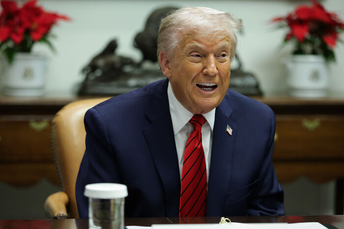 Former President Trump sitting at a table indoors, smiling and wearing a navy suit with red striped tie. Former President Trump sitting at a table indoors, smiling and wearing a navy suit with red striped tie.