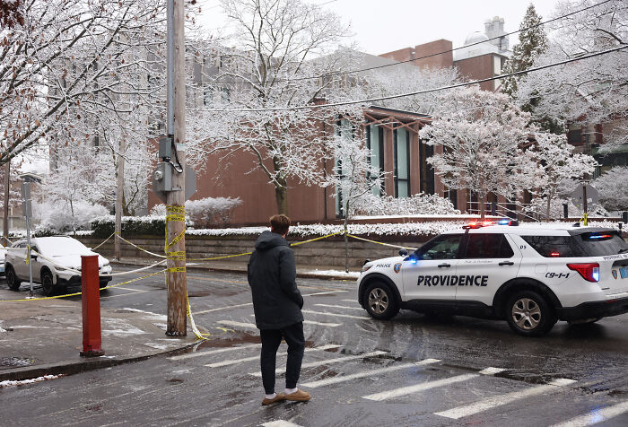 Police car with flashing lights blocking snowy street at a crime scene with caution tape in an urban area.