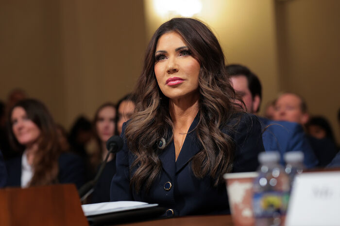 Kristi Noem testifying at a hearing, dressed in dark blazer, with focused expression and audience in background. Kristi Noem testifying at a hearing, dressed in dark blazer, with focused expression and audience in background.