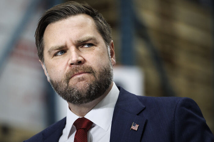 JD Vance in a dark suit and red tie, looking serious while attending a public event indoors.