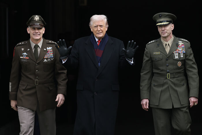 Donald Trump standing between two military officers, wearing a dark coat and gloves, related to Trump’s war crimes in the Caribbean. Donald Trump standing between two military officers, wearing a dark coat and gloves, related to Trump’s war crimes in the Caribbean.