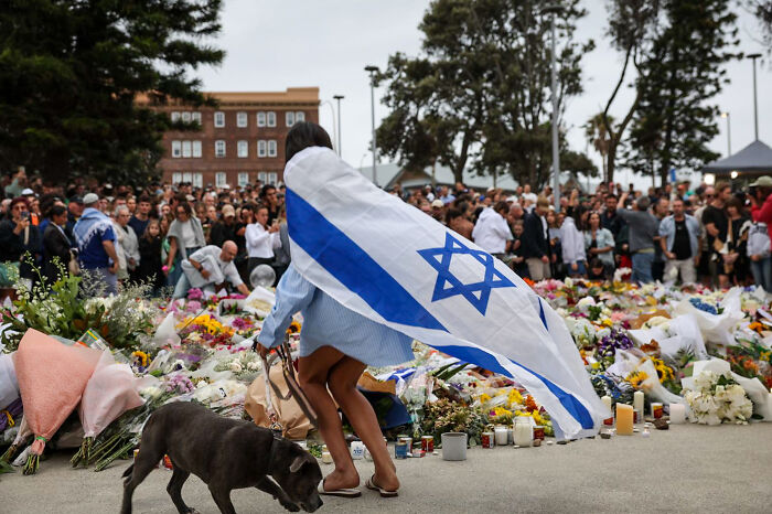 Woman draped in Israeli flag at Bondi Beach memorial with crowd honoring victims after Hanukkah attack gunman incident. Woman draped in Israeli flag at Bondi Beach memorial with crowd honoring victims after Hanukkah attack gunman incident.