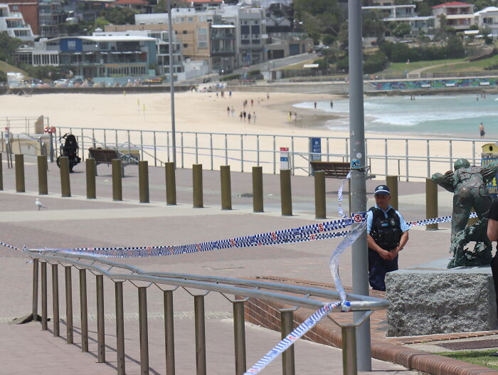 Police officer stands near taped-off area at Australian beach promenade amid tightened gun laws after mass shooting debate. Police officer stands near taped-off area at Australian beach promenade amid tightened gun laws after mass shooting debate.