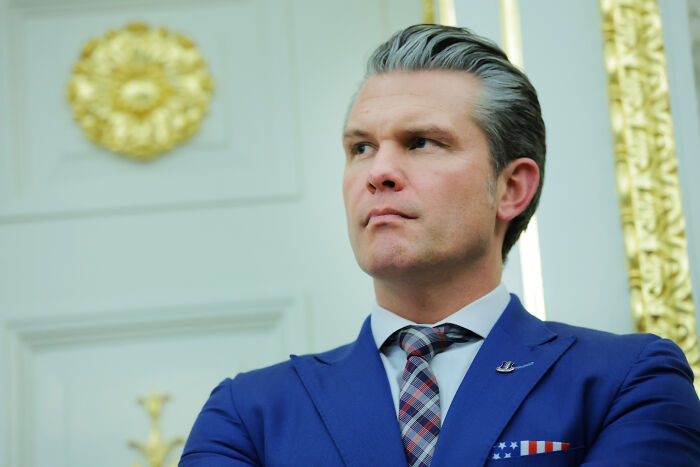 Man in a blue suit with an American flag pocket square, standing in a formal room with gold accents and decorations Man in a blue suit with an American flag pocket square, standing in a formal room with gold accents and decorations