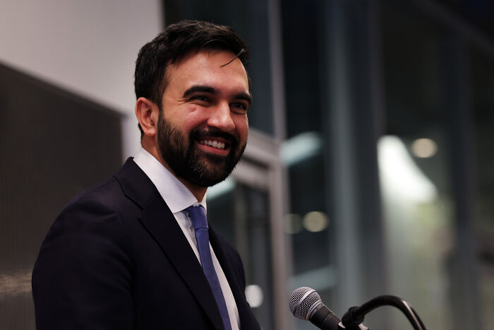 Smiling man in a suit speaking at a microphone during an indoor event related to Kim Petras and Zohran Mamdani. Smiling man in a suit speaking at a microphone during an indoor event related to Kim Petras and Zohran Mamdani.