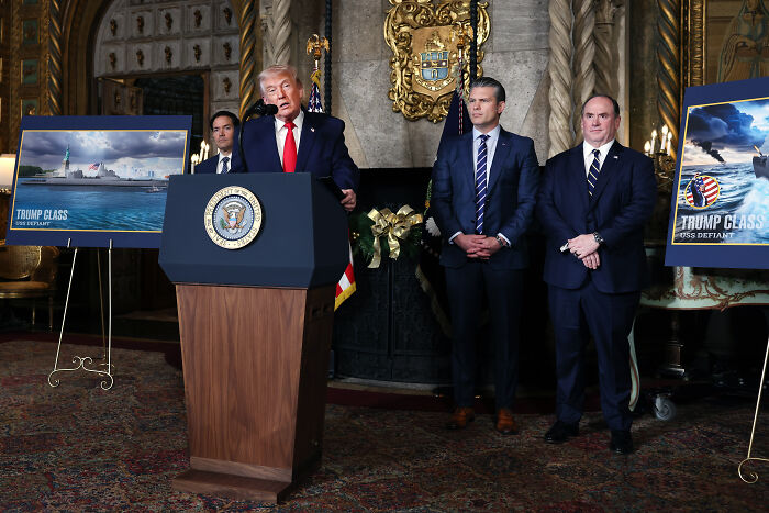 Donald Trump speaking at a podium unveiling new battleships named after his favorite president with aides standing nearby.