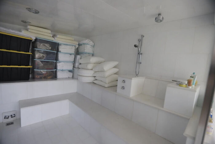 Minimalist white tiled bathroom with stacked pillows, plastic storage bins, and a showerhead on the wall.