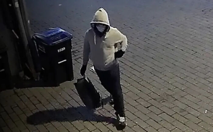Masked individual in a light-colored hoodie carrying a black case on a paved street in DC pipe b**b investigation footage.
