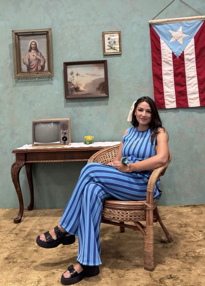 AOC seated indoors near Puerto Rico flag, vintage TV, and paintings, relating to luxury hotels and Puerto Rico venue visit. AOC seated indoors near Puerto Rico flag, vintage TV, and paintings, relating to luxury hotels and Puerto Rico venue visit.