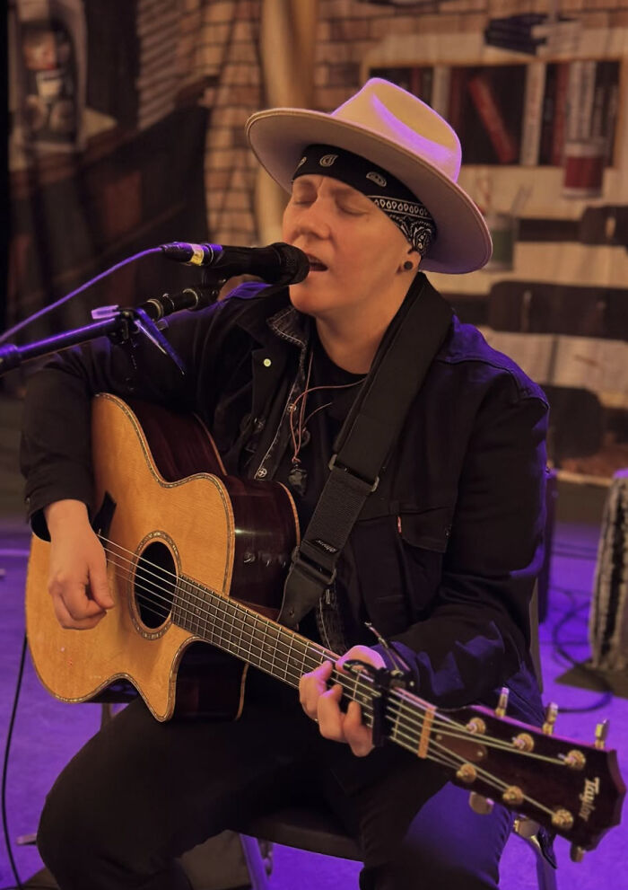Musician wearing a hat and bandana playing acoustic guitar and singing into microphone during a live performance.